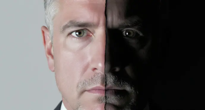 A close-up, half-lit portrait of a mixed-light-skinned MIDDLE AGED PROFESSIONAL face. One side is brightly lit and shows a confident, commanding expression. The other side is in shadow, revealing a subtle, almost imperceptible look of profound weariness. This image is a powerful metaphor for the hidden emotional cost of leadership.